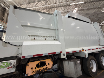 Truck side view showing a white Peterbilt 520 garbage truck body with steel hopper and dual axles in an indoor facility.