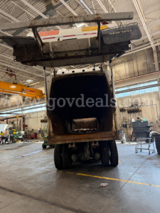 Peterbilt 520 rear view with tailgate and waste compactor bin raised, inside a shop. VIN label not visible.