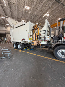 Peterbilt 520 truck in a maintenance bay with a large white refuse body mounted on a trailer; various labels and hydraulic components visible on the rear and side doors.