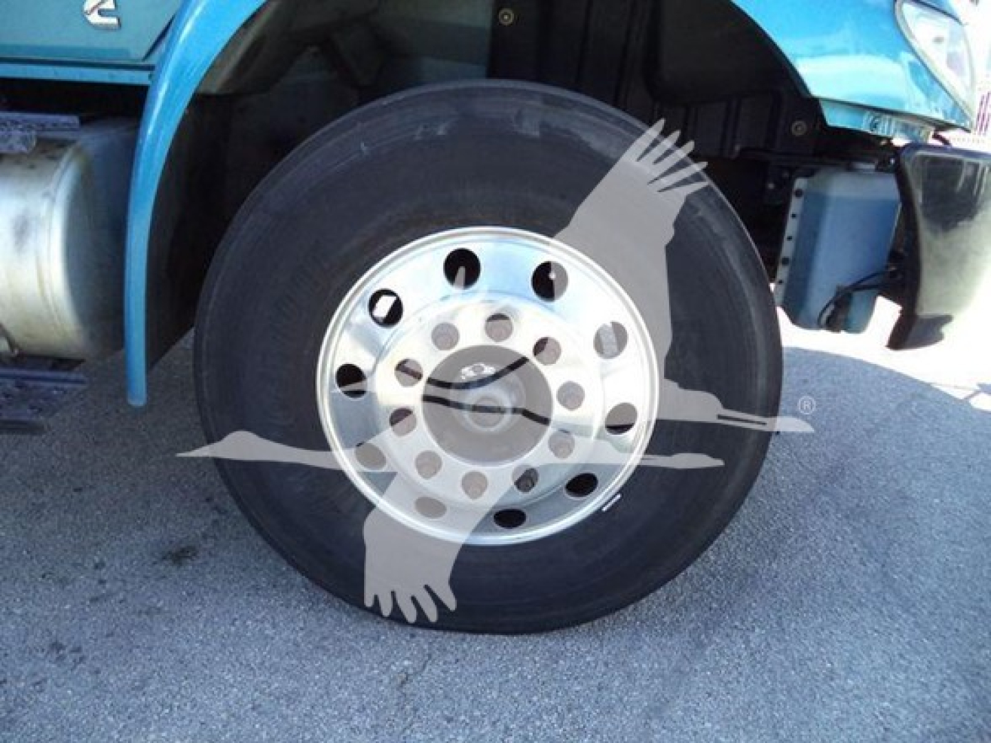 Truck wheel with large black tire and silver multi-hole hubcap under the fender of a blue International MV dump truck.