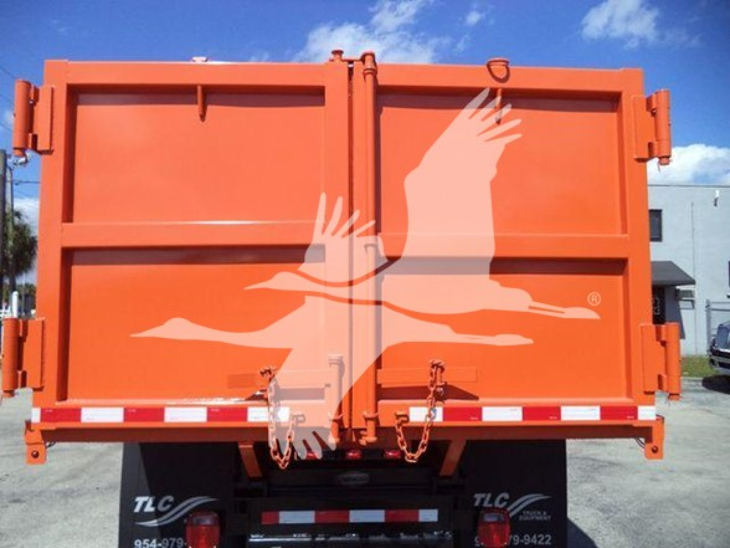 Orange dump truck tailgate with hydraulic latches and chain hooks, TLC logo visible on lower left and right bumper area.