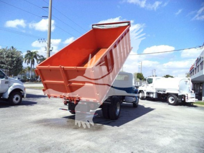 Dump truck bed tilted up showing cargo bed and hydraulic mechanism under the orange bed with white cab trucks in background.