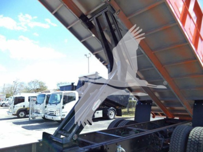 Dump truck bed raised with hydraulic cylinder visible; front cab area and other trucks in lot; side of bed shows mechanical linkage and chassis components.
