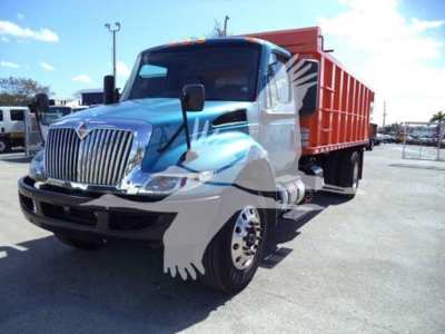 Truck in a lot showing a blue International MV dump truck with an orange dump body. Front grille, large mirrors, and headlights visible.