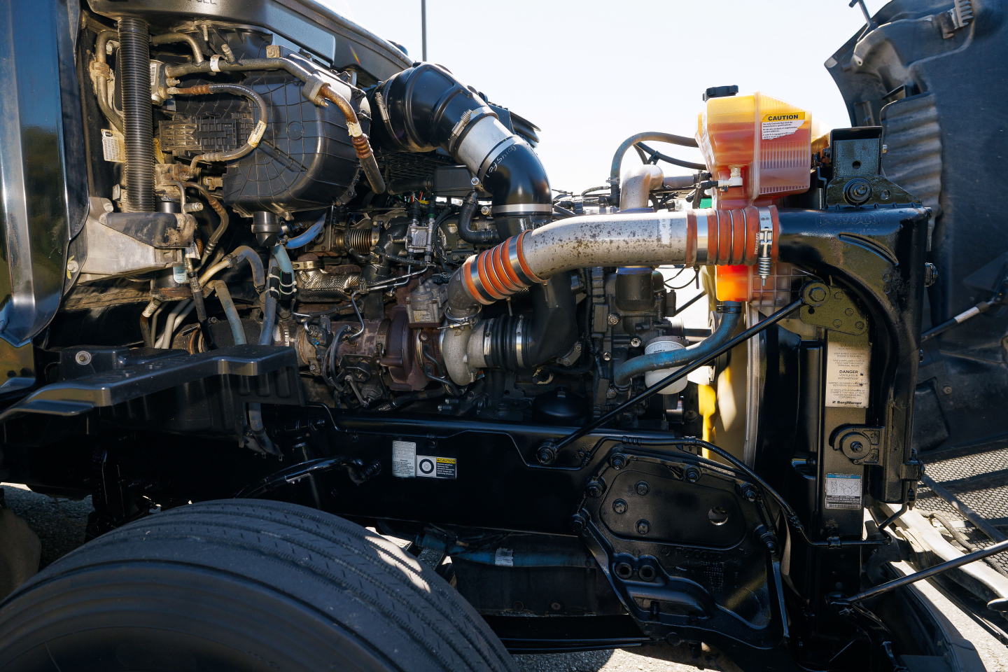 Peterbilt 579 engine bay showing turbocharger, intake piping, orange coolant reservoir, and various hoses and components.