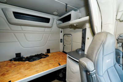 Peterbilt cab interior of a 2019 579 sleeper, showing the bunk area with plywood bed surface, overhead storage shelf, and driver seat in the foreground.