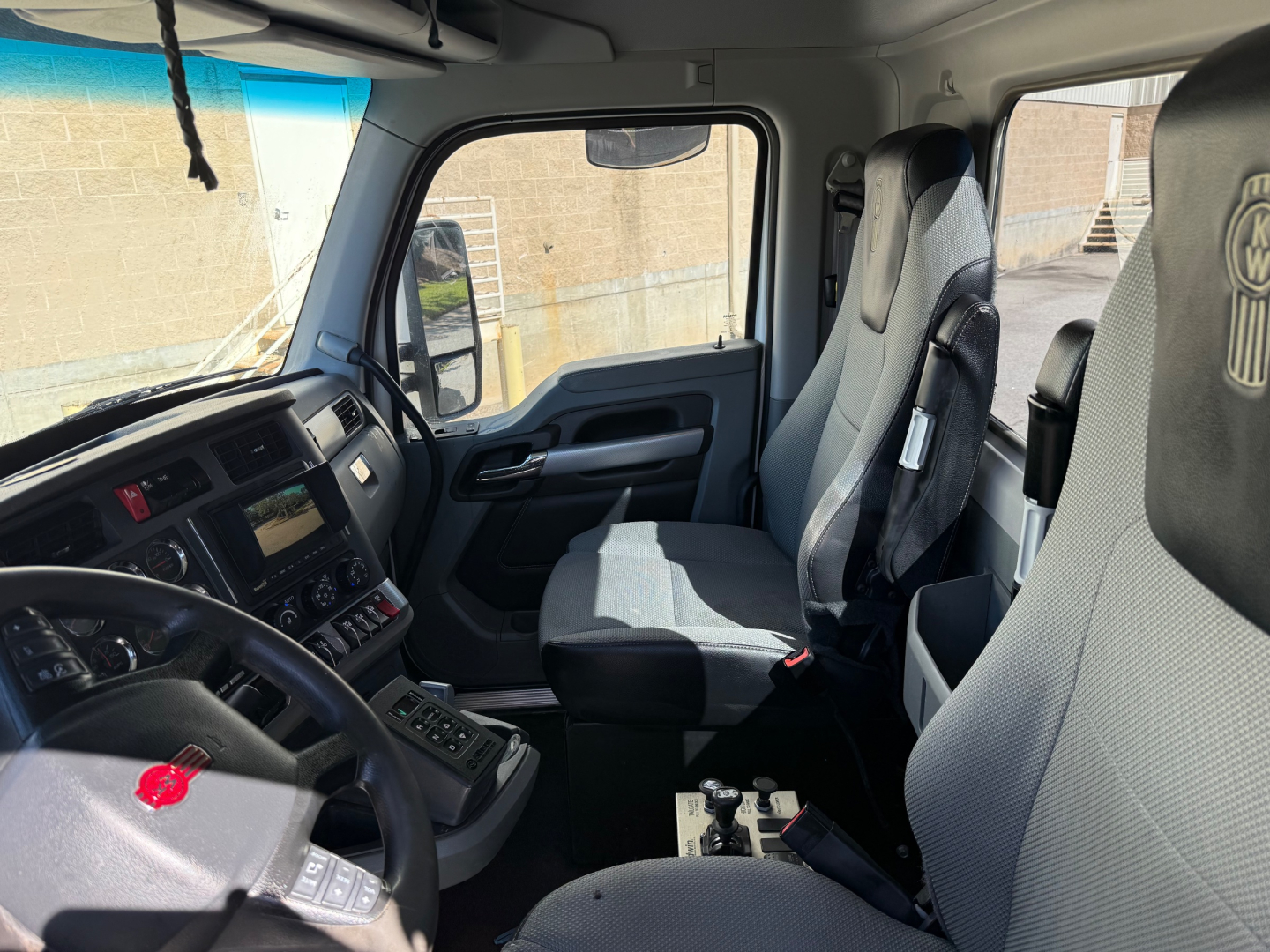 Kenworth T880 truck cabin interior showing steering wheel, dashboard with controls, driver seat, and passenger seat. Glass windows and door with side mirror visible.