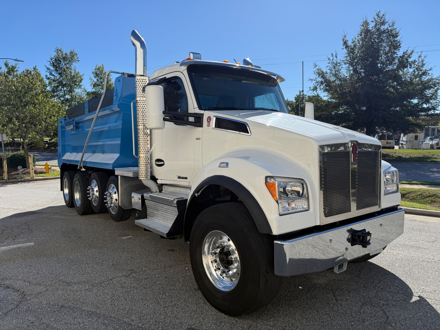Kenworth T880 dump truck in white cab with blue dump body, large grille, side exhaust stacks, and dual rear wheels. Visible emblem and polished chrome bumper.