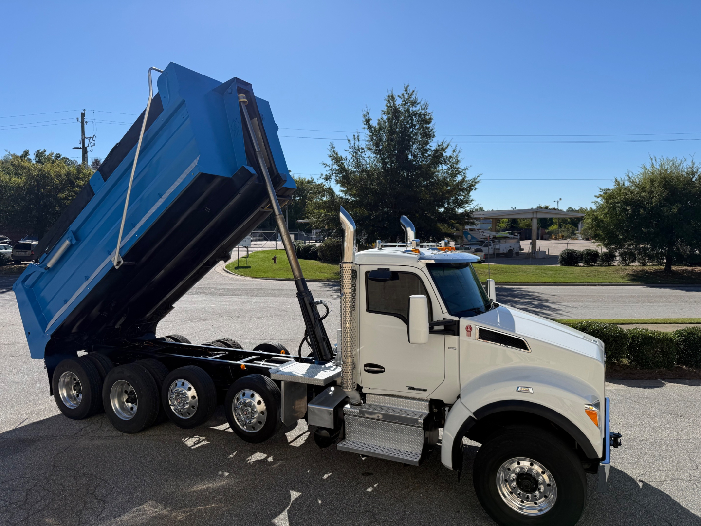 Kenworth T880 dump truck with blue bed raised on a white cab. Side shows multiple wheels and chrome fuel tanks; no legible labels visible.