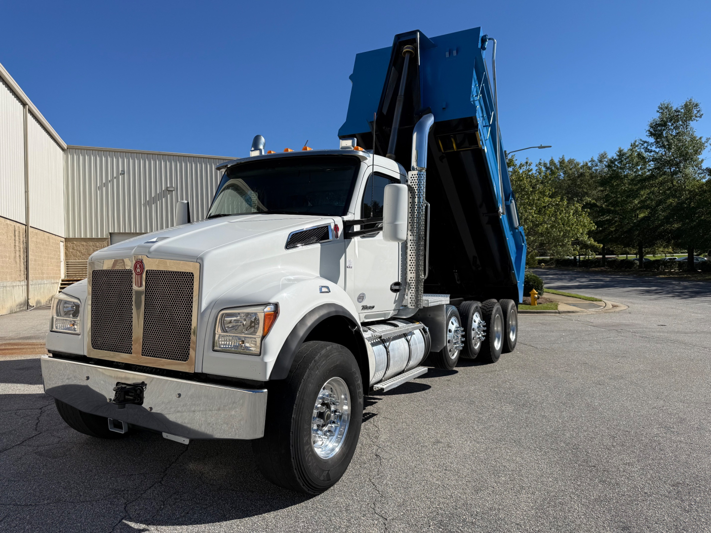 Kenworth T880 dump truck in white cab with a blue dumping bed raised, side view showing large grille, dual rear axles, chrome exhaust stacks.