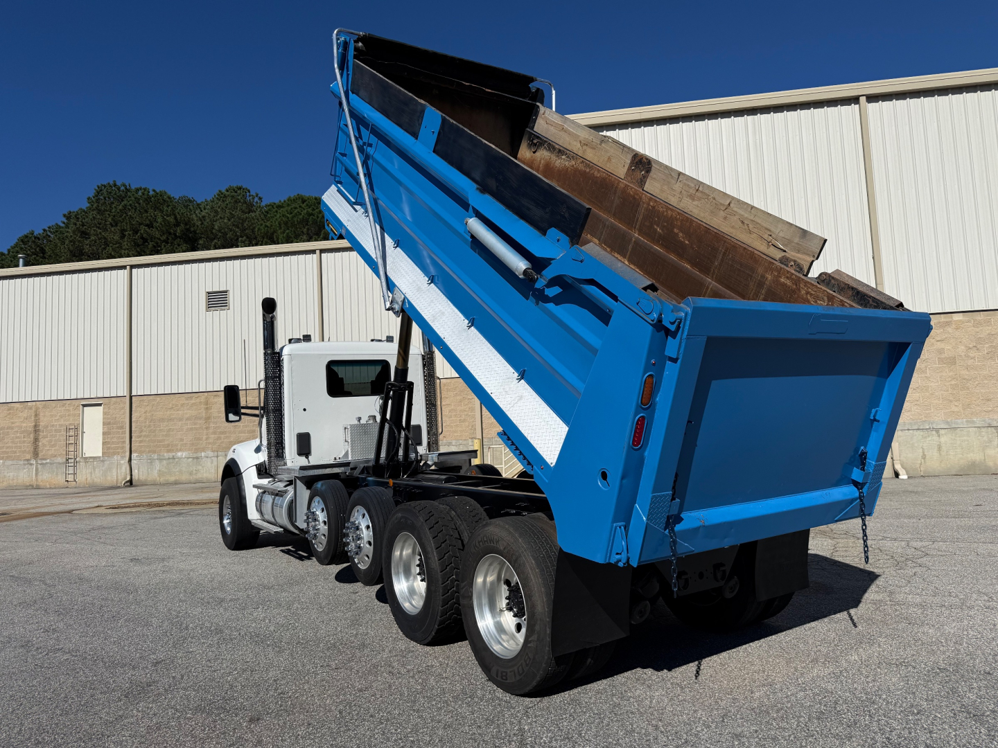 Kenworth T880 dump truck in raised bed showing rear view with blue hull and loading bed, three-axle configuration, white cab, dealership lot.
