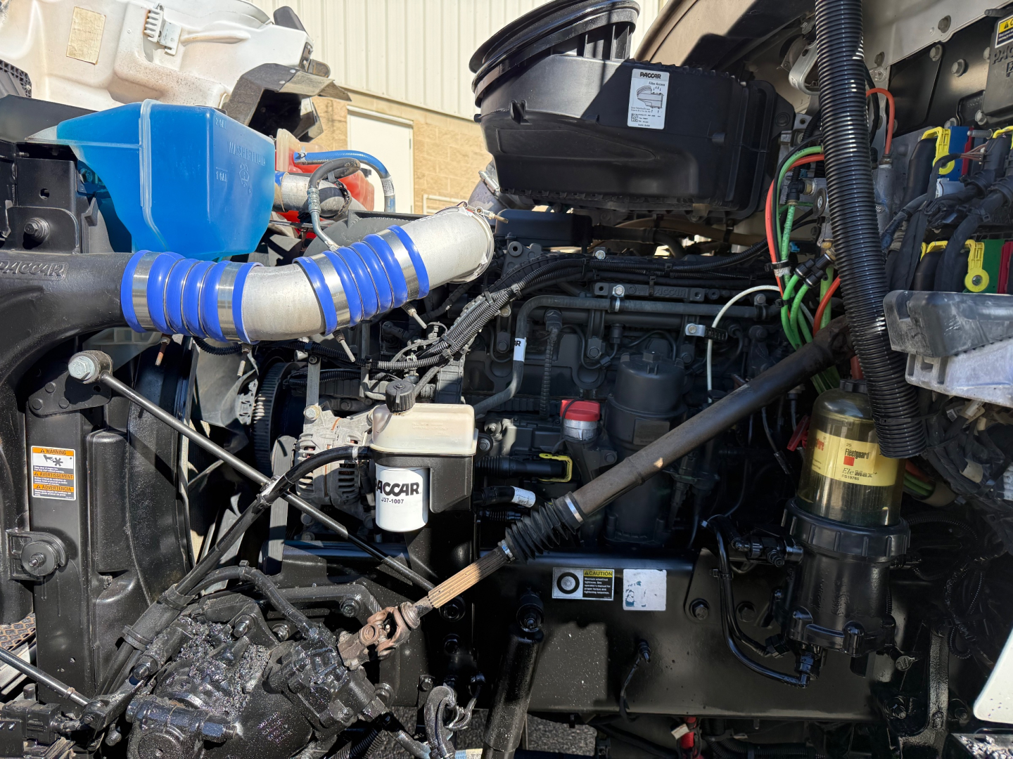 Kenworth T880 engine bay showing intake hoses, air filter housing, coolant reservoir, and various hoses and wiring. Visible components include turbo/intercooler piping, oil filter, and engine block details.