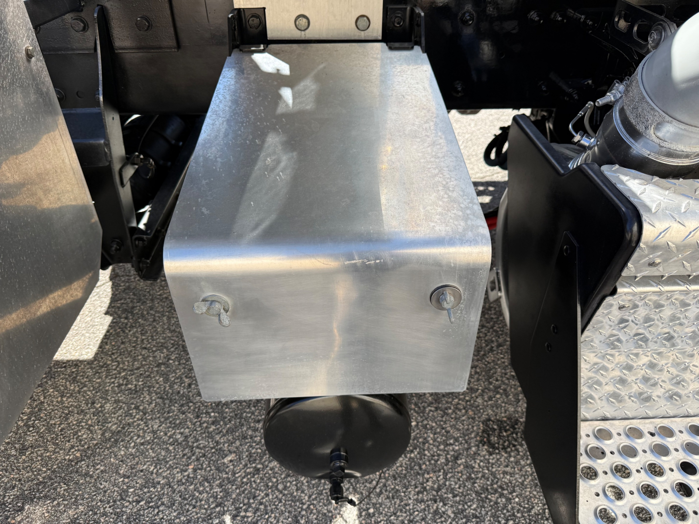Truck component: metal aluminum toolbox or fuel tank mounted under the chassis between rear axles, with mounting bolts and a black circular connection below; surrounding metal steps and chassis visible.