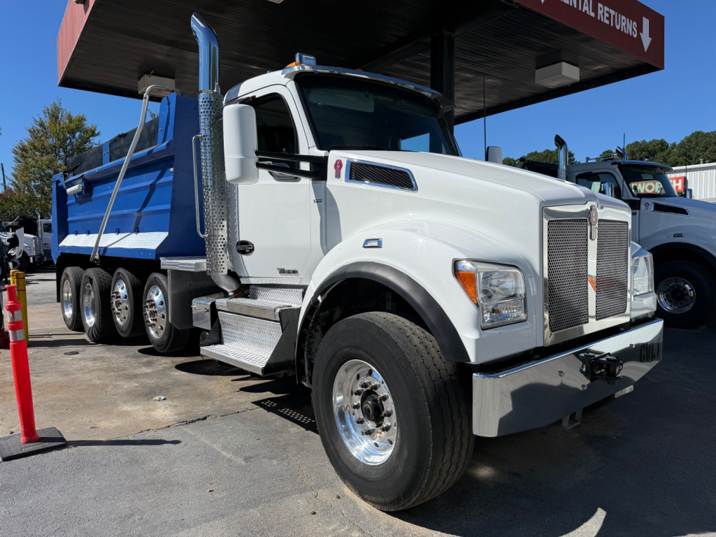 Kenworth T880 dump truck in a dealership lot, front-left view, white cab with chrome grille and side exhaust, blue dump bed, multiple axles.