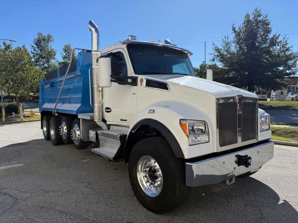 Kenworth T880 dump truck in white cab with blue dump body, large grille, side exhaust stacks, and dual rear wheels. Visible emblem and polished chrome bumper.