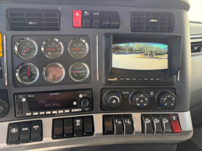 Kenworth T880 instrument cluster and dashboard view showing multiple gauges, orange hazard switch, emergency controls, a small monitor, and radio/display panel.