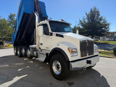 Kenworth T880 dump truck in white with blue dump bed raised, front view shows large grille, dual exhaust stacks, chrome bumper, and heavy-duty tires.