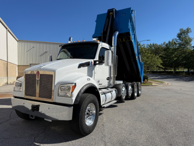 Kenworth T880 dump truck in white cab with a blue dumping bed raised, side view showing large grille, dual rear axles, chrome exhaust stacks.