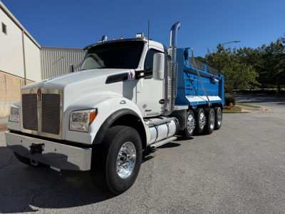 Kenworth T880 dump truck front-left view with blue dump bed and chrome wheels, diesel exhaust stacks, daytime outdoor lot.