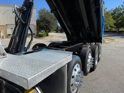 Kenworth T880 dump truck rear undercarriage showing dual rear axles with chrome wheels and raised dump bed; hydraulic lifting mechanism and diamond plate toolbox visible.