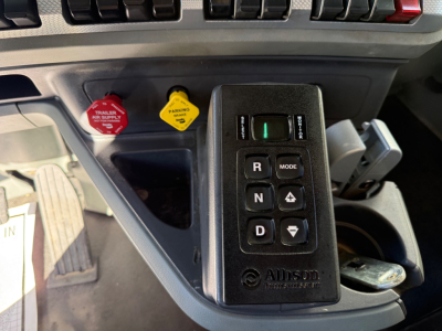 Kenworth T880 interior with joystick shift module showing R N D P and mode buttons; yellow parking brake and red trailer air supply knobs nearby.
