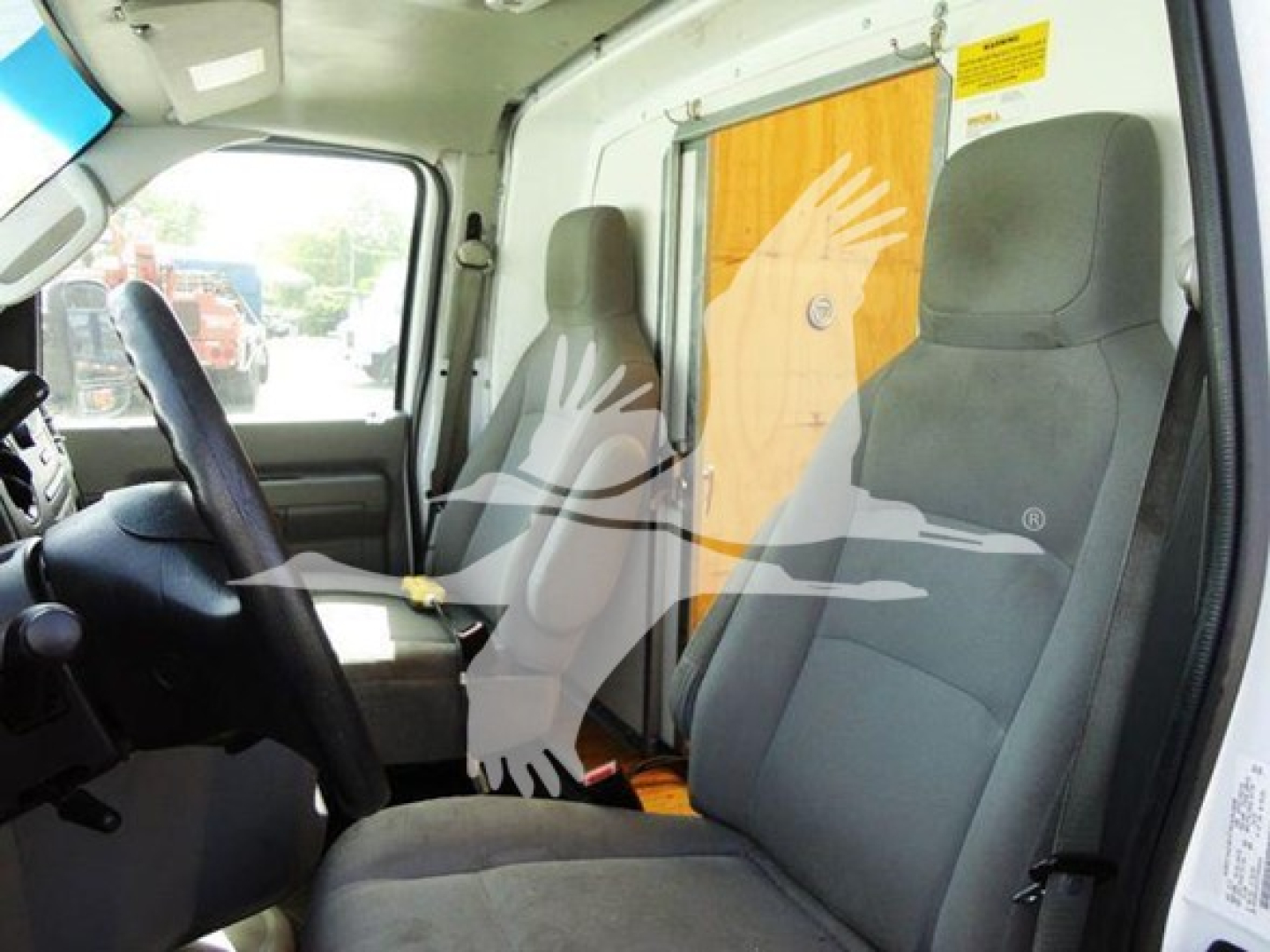 Truck interior: driver’s seat and passenger seat with steering wheel, dashboard, and a partition wall with a wooden panel behind. Visible labels near top window.