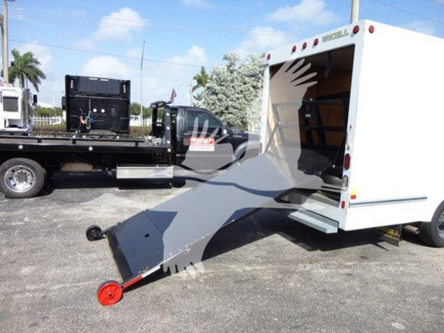 Ford E-350 cutaway van with a white rear cargo area and a metal loading ramp extended to the ground; a flatbed tow truck is in the background.