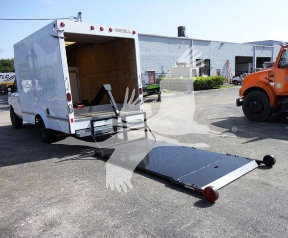 Ford E350 box truck with a lowered rear ramp and open cargo area, white high-roof body, black ramp plate, and orange tow truck in background.