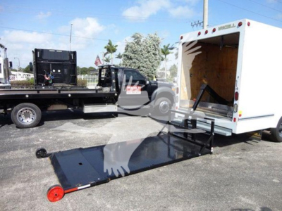 Ford E350 Econoline cutaway truck with a lowered wheelchair/roll-off ramp next to a white cargo box; rear access doors open showing interior plywood walls.