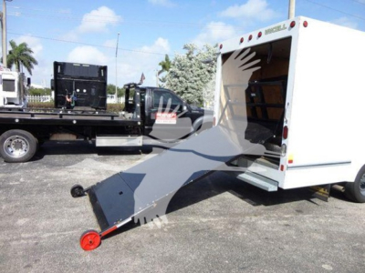 Ford E-350 cutaway van with a white rear cargo area and a metal loading ramp extended to the ground; a flatbed tow truck is in the background.