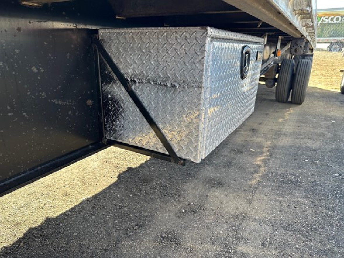 Underbelly view of a utility spread air combo flatbed showing a diamond-plate aluminum toolbox under the trailer side with clamps and wheels in background.