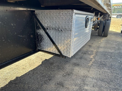 Underbelly view of a utility spread air combo flatbed showing a diamond-plate aluminum toolbox under the trailer side with clamps and wheels in background.