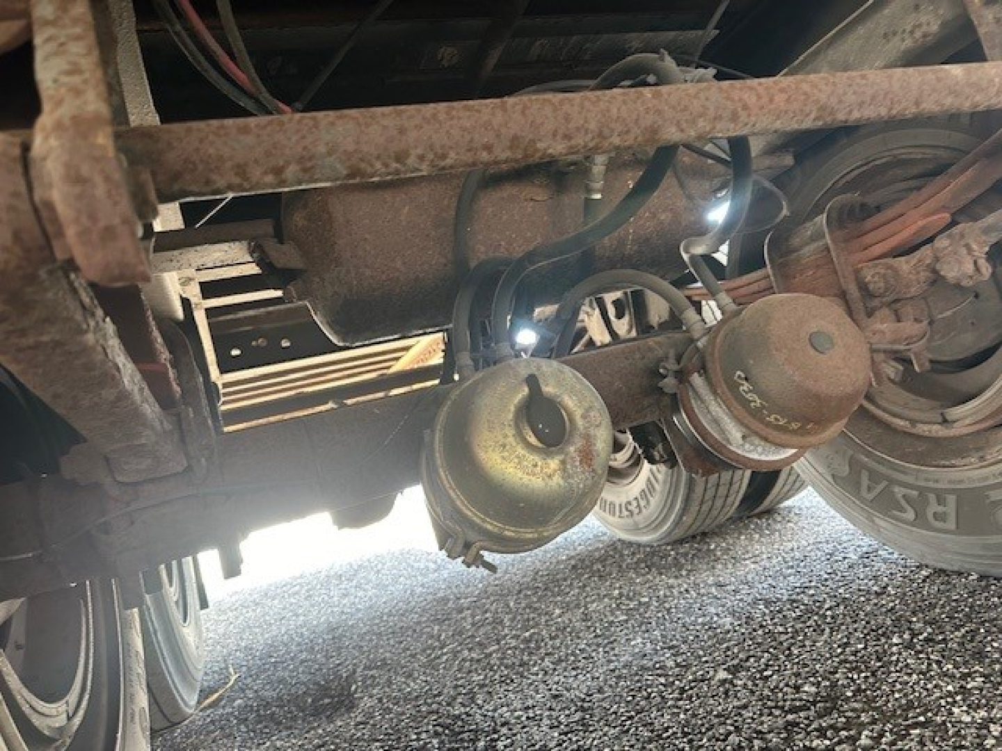 Trailer undercarriage with twin leaf springs, brake chambers, and plumbing; rusted frame visible; rear wheel and tire in view.
