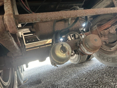 Trailer undercarriage with twin leaf springs, brake chambers, and plumbing; rusted frame visible; rear wheel and tire in view.