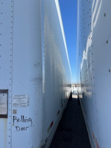 Trailer side walls of a stacked roll door trailer showing close gap between two white trailers with a narrow driveway; handwritten text 'Rolling Door' on the left trailer.