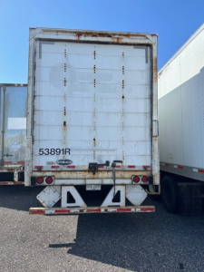 Rear view of a weathered Strick roll door trailer showing worn metal panels, rust, and Strick logo plate near lower left of door.