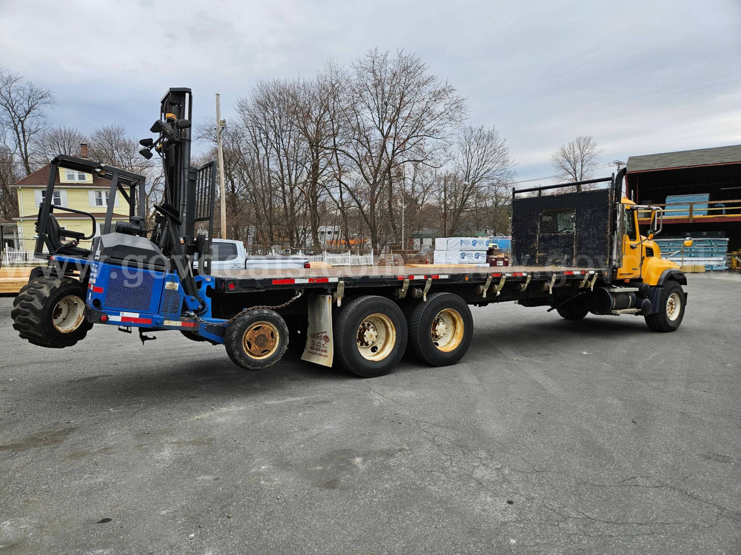 Mack CV713 lowboy tractor with flatbed trailer and mounted forklift system on rear, side view showing long multi-axle trailer and yellow tractor unit.
