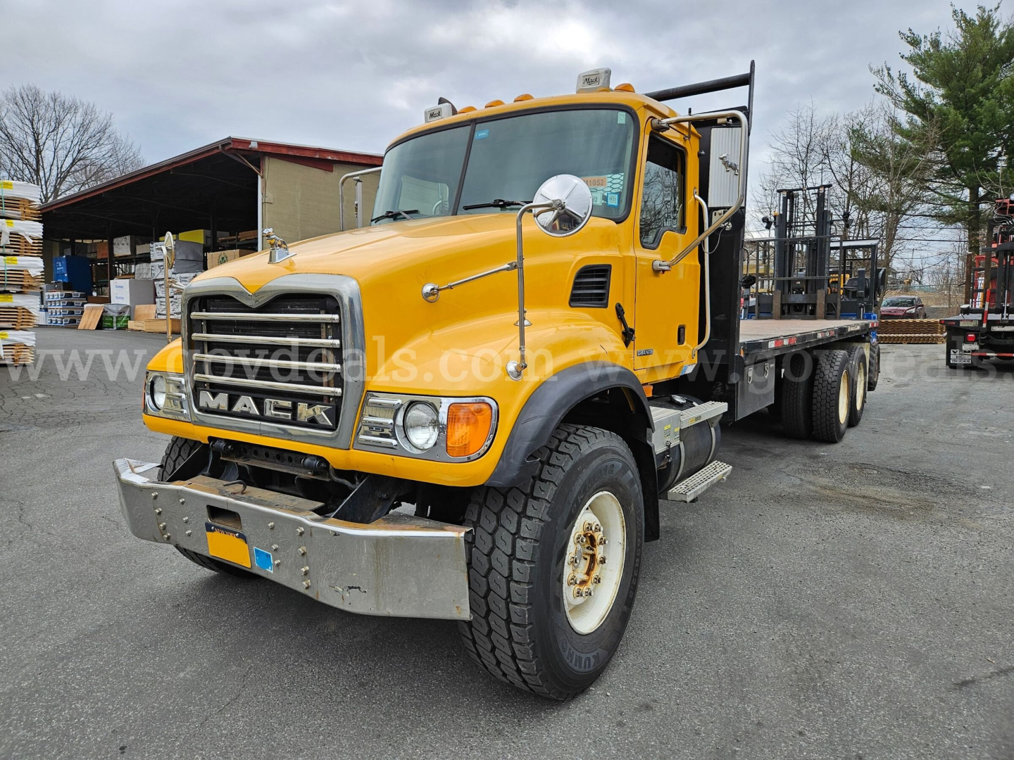 Yellow 2005 Mack CV713 flatbed truck in three-quarter front view showing large grille, bumper, dual headlights, and side mirrors.