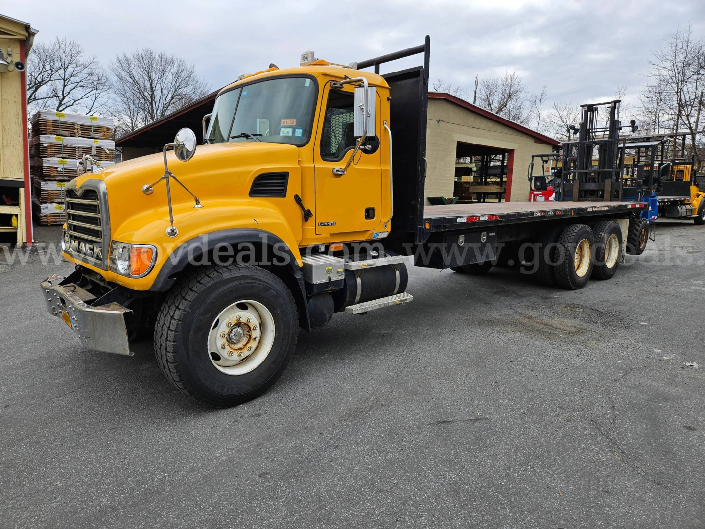 Mack CV713 flatbed truck in yellow, side view showing front grille, cab, and long flatbed with tandem rear axles.