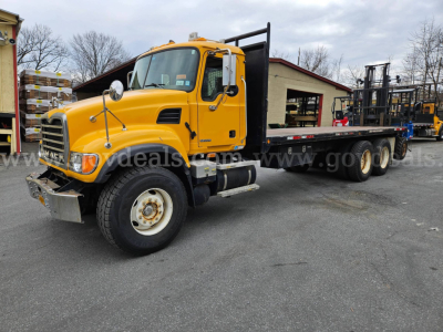 Mack CV713 flatbed truck in yellow, side view showing front grille, cab, and long flatbed with tandem rear axles.