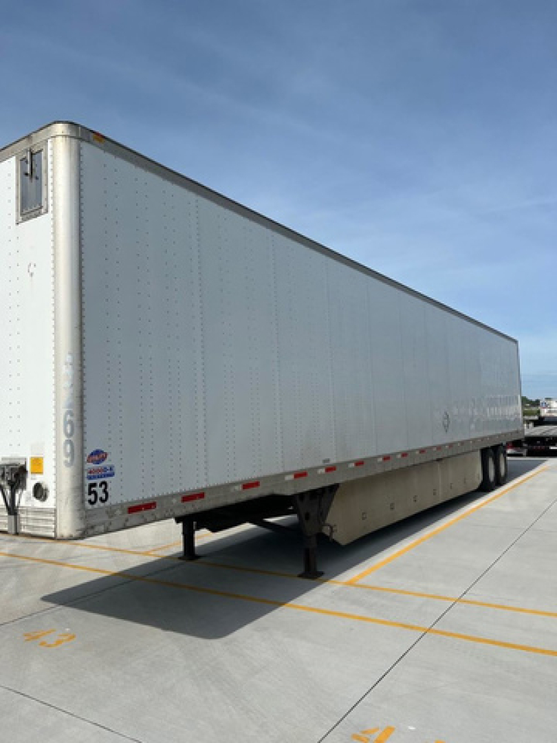 Trailer with box body and landing gear visible. Side shows riveted aluminum exterior with small logo near rear, front corner with wheel well, and numbered markings near bottom left. Clear blue sky.