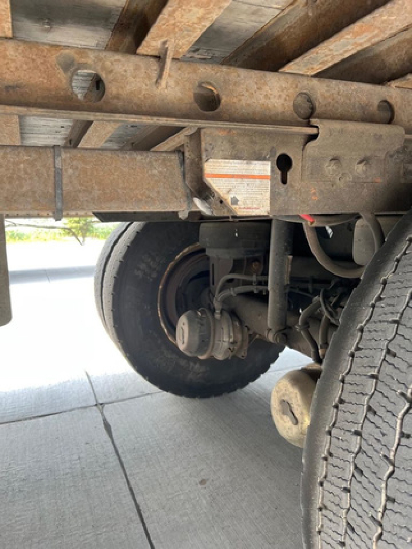 Trailer undercarriage with suspension, axle, and wheel assembly visible; rust on frame; warning label on crossmember; tire tread visible.