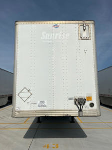 Box trailer rear view showing large white Door with small top vent, utility van trailer details, VIN label, and electrical plug area.