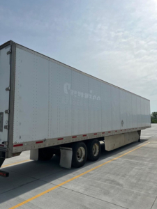 Trailer near side view showing a large white box trailer with triple axle and undercarriage; some faded markings on the side.