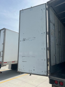 Rear cargo area of a utility van trailer with open door showing interior metallic walls and rivets.