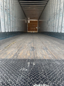 Inside view of an empty utility van trailer showing wooden floor, metal sidewalls, and rear ramp door.