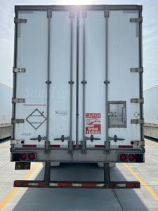 Rear view of a light-colored utility van trailer with double doors, caution sticker, and taillights.