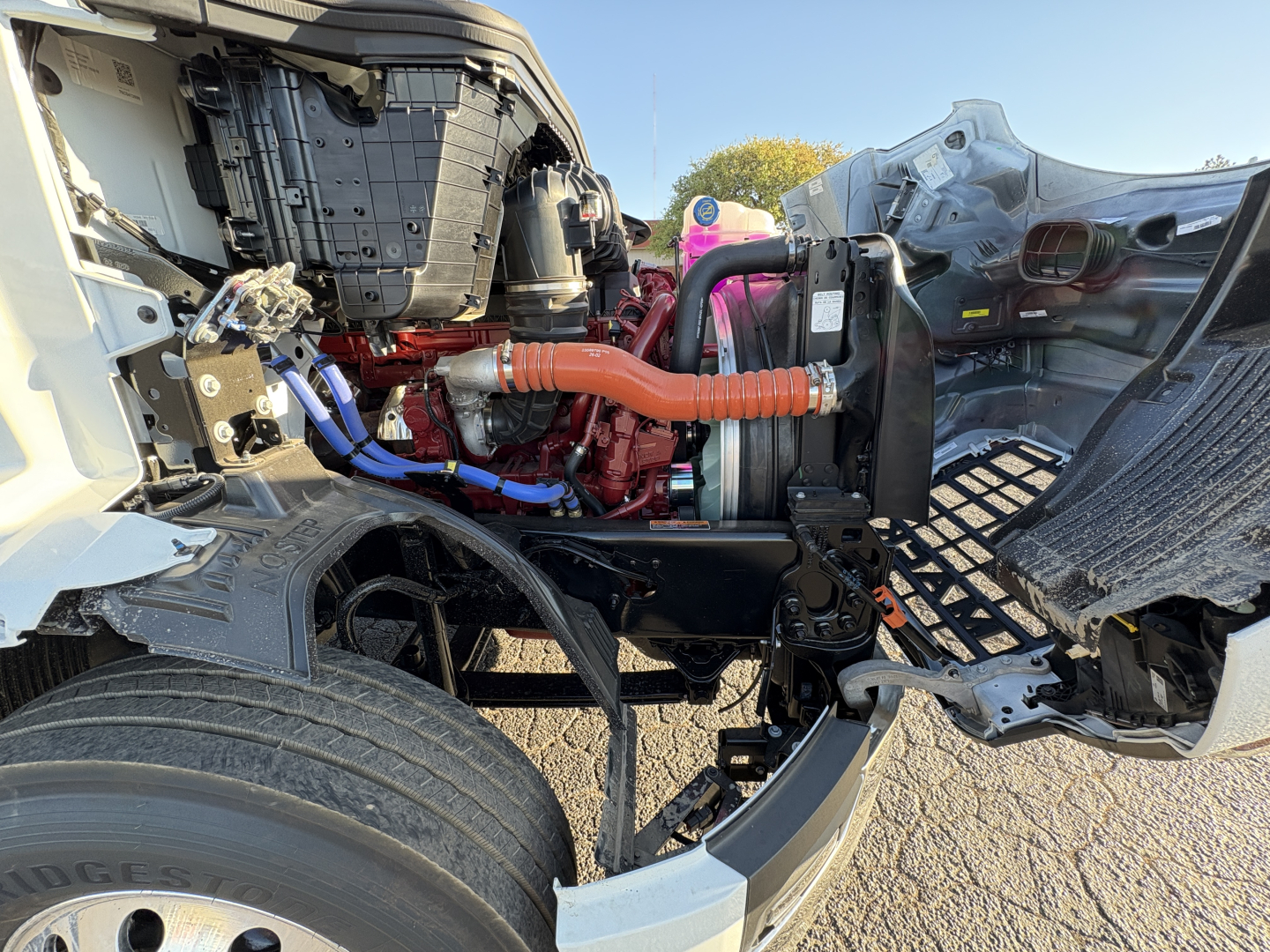 Engine bay of a 2027 Mack AN64T conventional daycab showing orange high-voltage hose, blue hoses, red engine block, radiator, and surrounding chassis components.