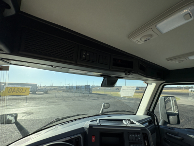 Truck cab interior showing overhead console with storage and controls, windshield view of a lot, side mirror, and dashboard of a 2027 Mack AN64T conventional daycab.