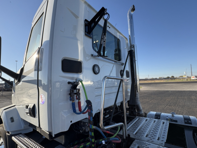 Front-left cab area of a 2027 Mack AN64T daycab showing external hoses and steps, with side mirrors and chrome exhaust stack visible.